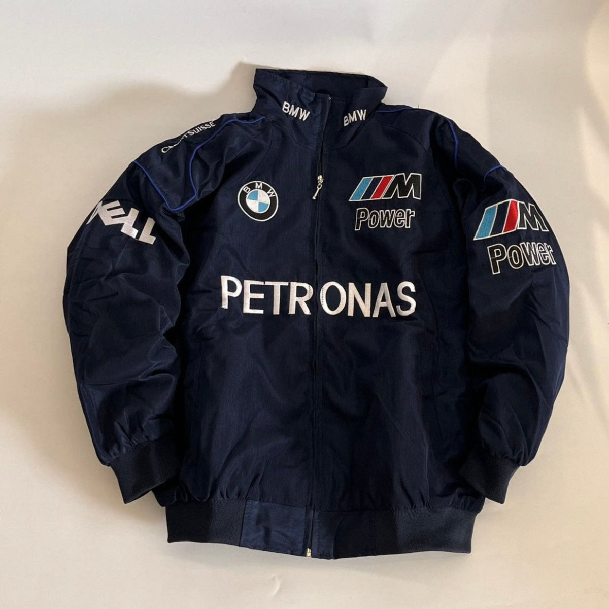 A navy blue BMW M2 Petronas racing jacket with white, blue, and red sponsor logos, laid flat on a light beige background.